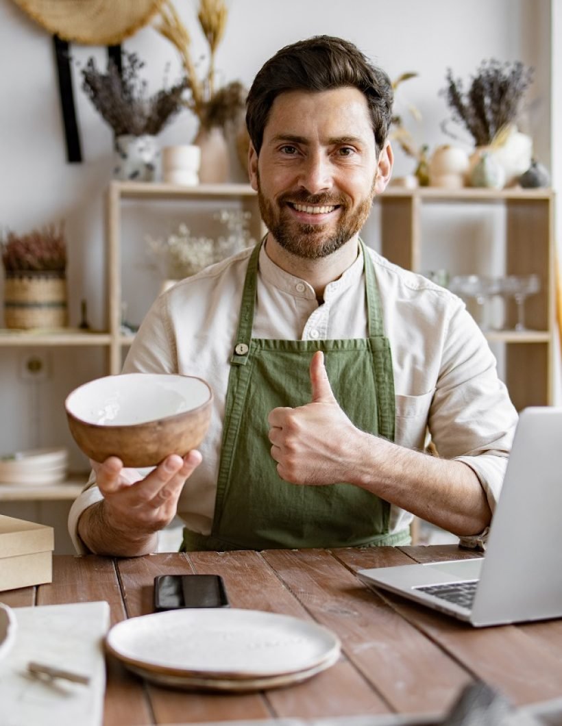 Happy worker in apron holds unique bowl created in personal style against background of shelves with dishes. Adult bearded male potter engaged in e-commerce selling handmade clay products on Internet.